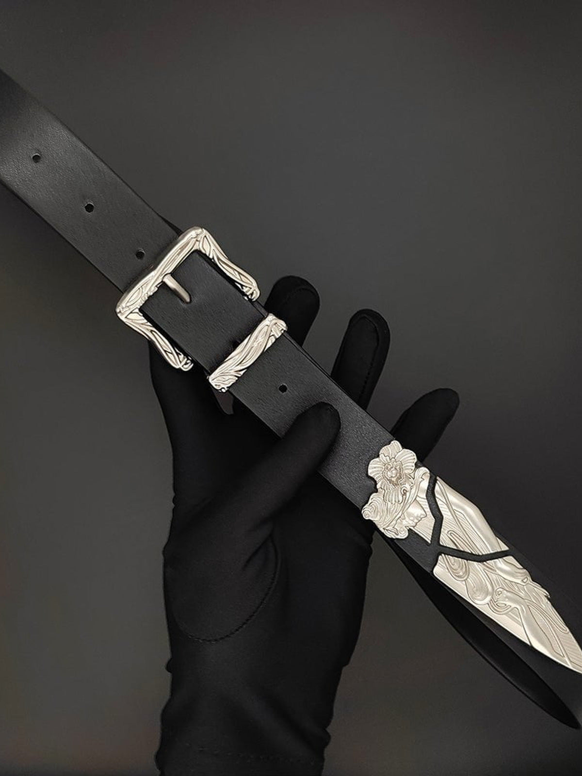Schwarzer Gürtel mit graviertem silberfarbenem Schnallen- und Spitzenbeschlag – in der behandschuhten Hand, Schnalle und Spitze | XoKool
